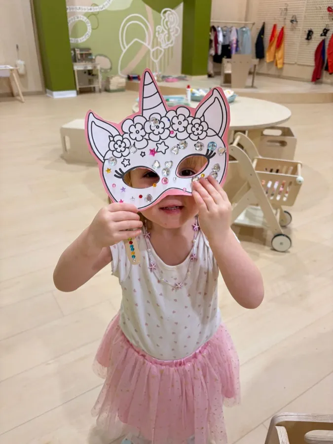A little girl in a tutu peeking through the unicorn mask she just decorated